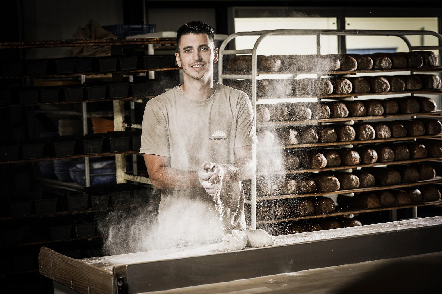 Unternehmer steht in seiner Backstube. Im Hintergrund sind Brote in Regalen. Auf der Arbeitsfläche liegt Teig. Das Mehl staubt.