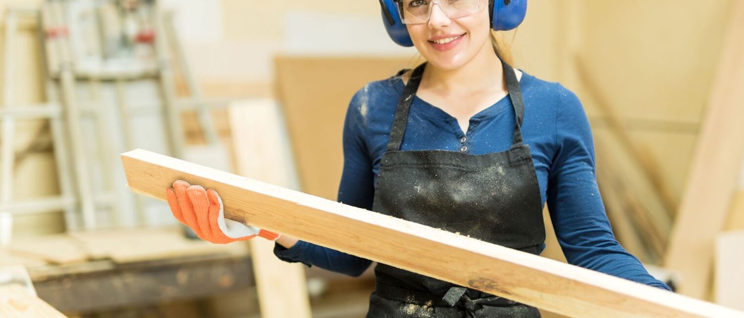 Junge Tischlerin in einer Werkstatt hält eine Holzplanke in den Händen und blickt in die Kamera.
