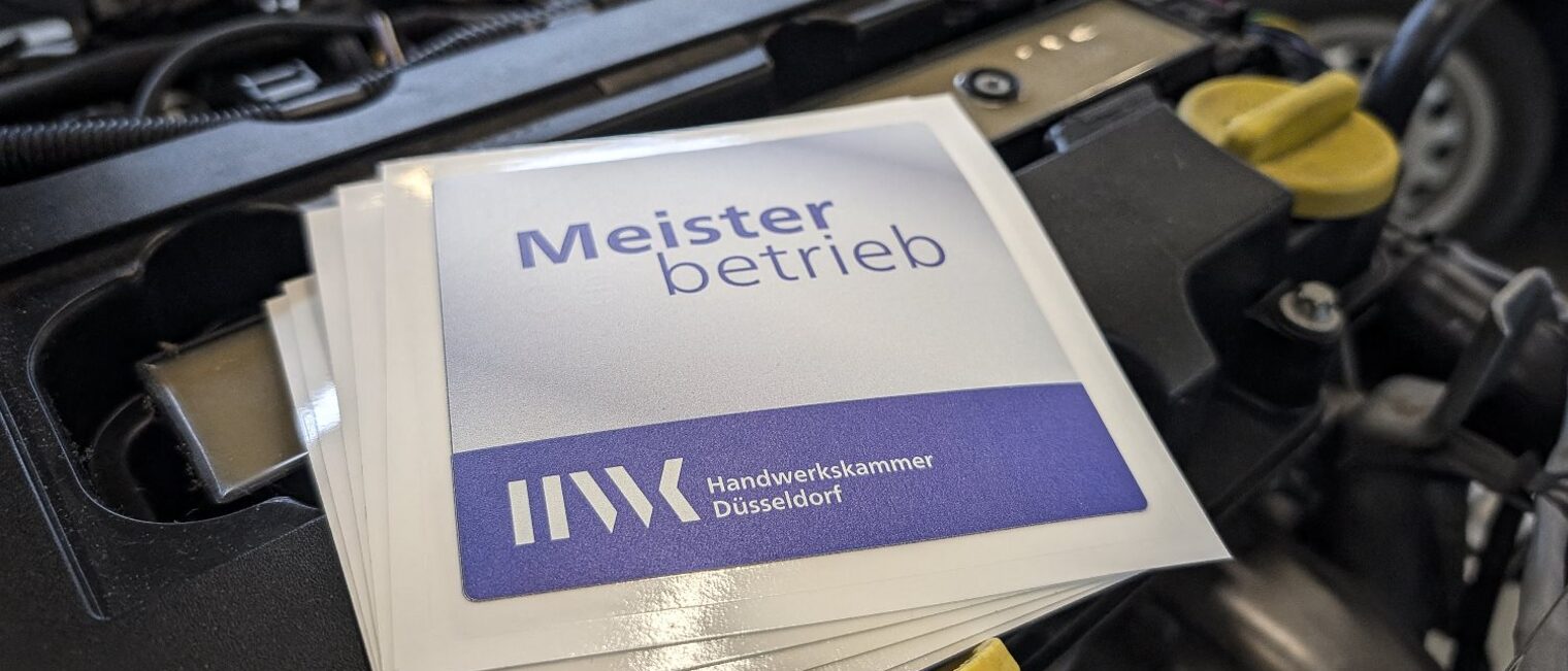 Mehrere Aufkleber mit Text "Meisterbetrieb" und Logo Handwerkskammer Düsseldorf liegen auf einem PKW-Motorblock.