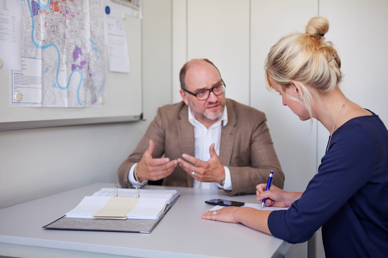 Beratungsgespräch zwischen einem Mann und einer Frau. Auf dem Tisch liegt ein geöffneter Ordner mit Unterlagen. Die Frau macht sich Notizen.